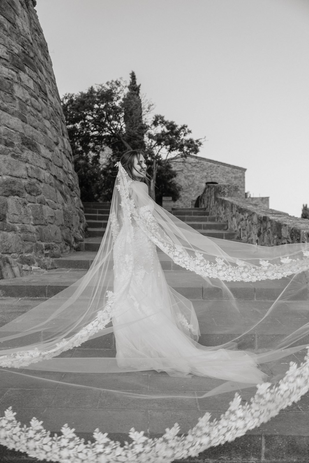 La núvia amb el vel al vent a les escales del castell, en blanc i negre